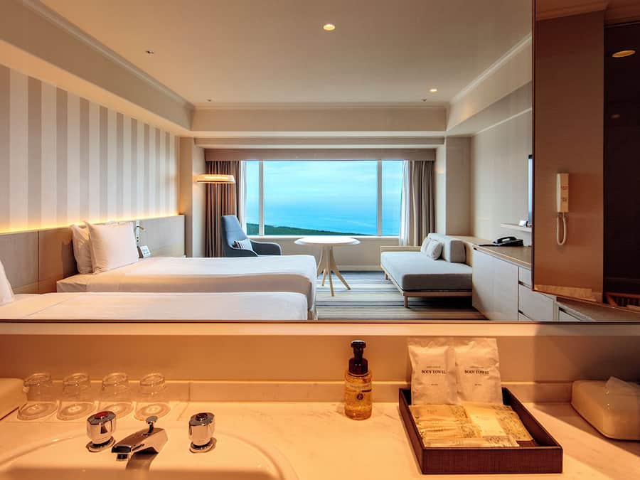 A hotel room with two beds, a sofa, a small table, a TV, and a bathroom with a sink and toiletries.