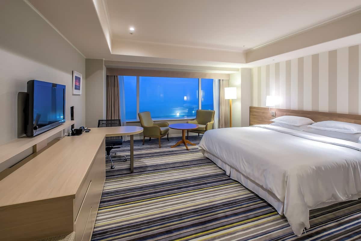 A hotel room with a large bed, a desk with a flat-screen TV, two chairs, a small round table, and a view of the ocean through large windows.