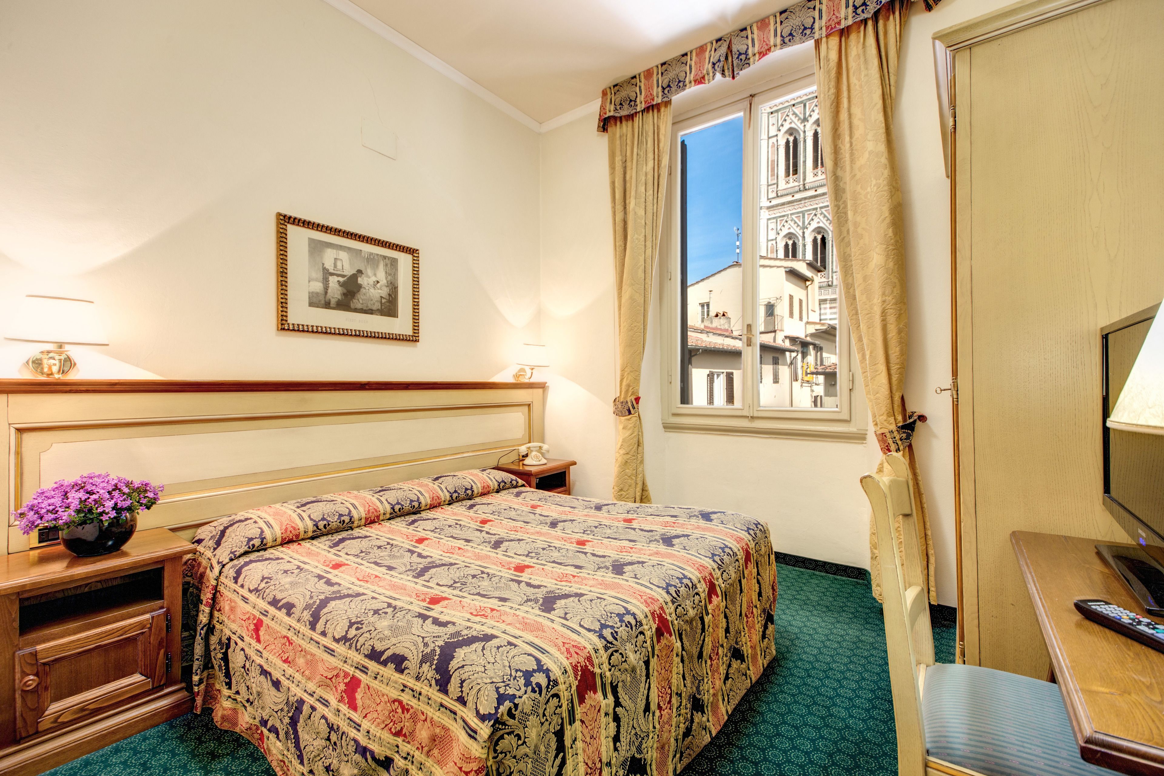 Une chambre d’hôtel avec un grand lit, une table de chevet avec une lampe, une petite table sur laquelle est posé un vase de fleurs, une chaise, un bureau avec un ordinateur et une fenêtre donnant sur un immeuble.
