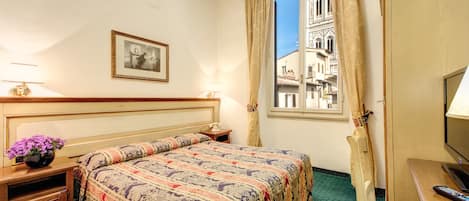 Une chambre d’hôtel avec un grand lit, une table de chevet avec une lampe, une petite table sur laquelle est posé un vase de fleurs, une chaise, un bureau avec un ordinateur et une fenêtre donnant sur un immeuble.