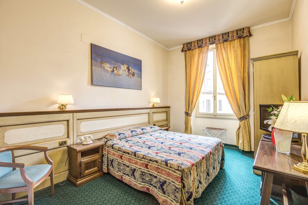 A hotel room with a bed, a chair, a desk, a lamp, a painting, and a window with curtains.
