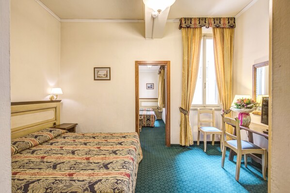 Habitación de hotel con una cama grande, un escritorio con silla, una ventana con cortinas y una puerta que da a otra habitación.