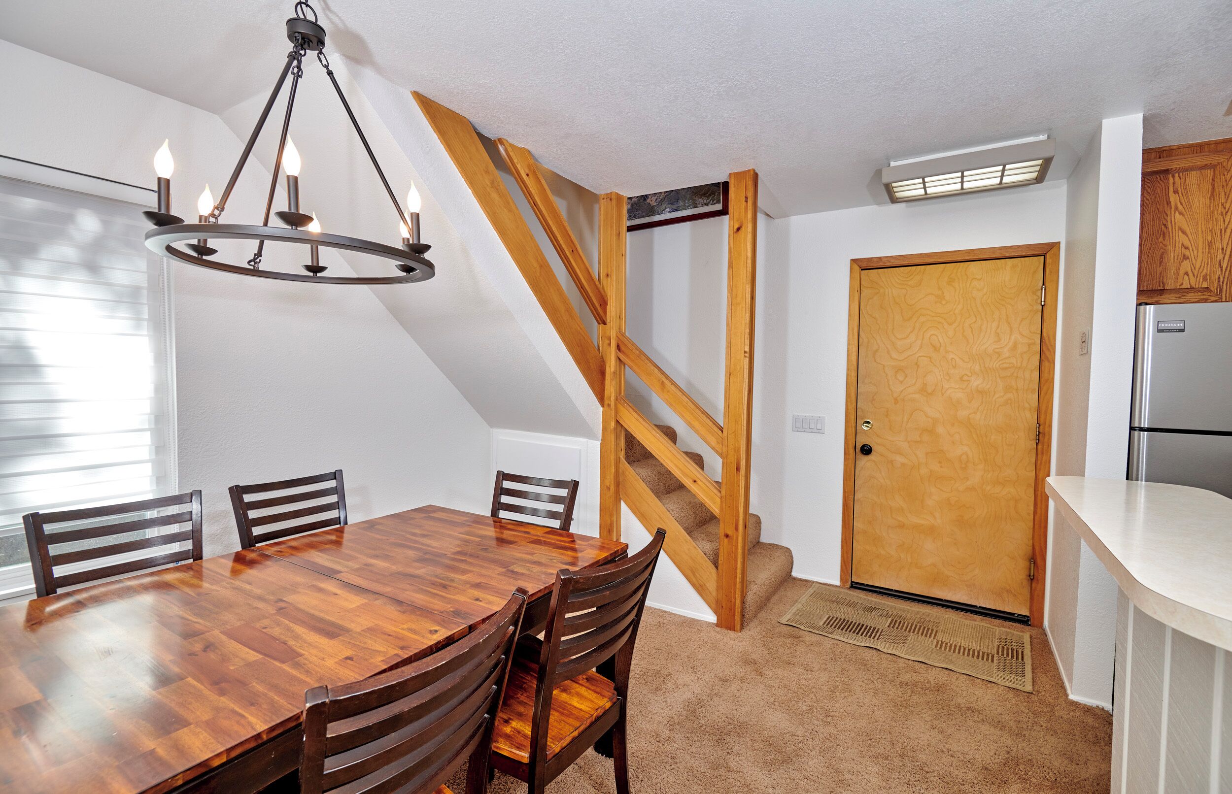 A dining area with a wooden table and chairs, a chandelier, a wooden staircase, and a kitchen area with a refrigerator.