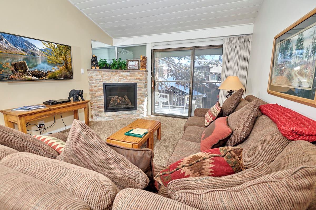 A cozy living room with a fireplace, a large window with a view of trees, and a comfortable sofa set.