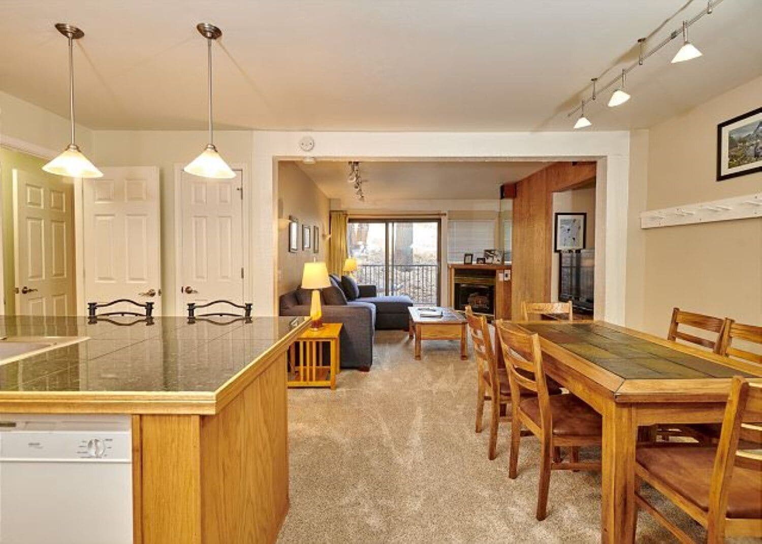 A modern kitchen with a central island, dining area with wooden chairs, and a living room with a sofa and lamp.