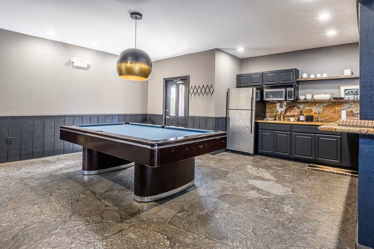 A pool table in a room with a kitchen area, a door, and a pendant light.