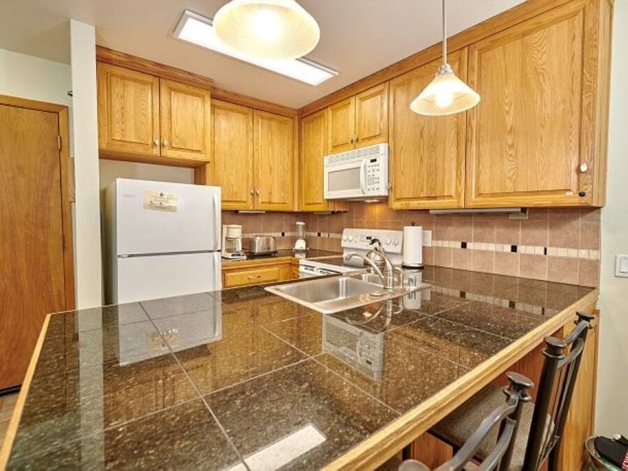 A kitchen with wooden cabinets, a stainless steel sink, a refrigerator, a microwave, and granite countertops.