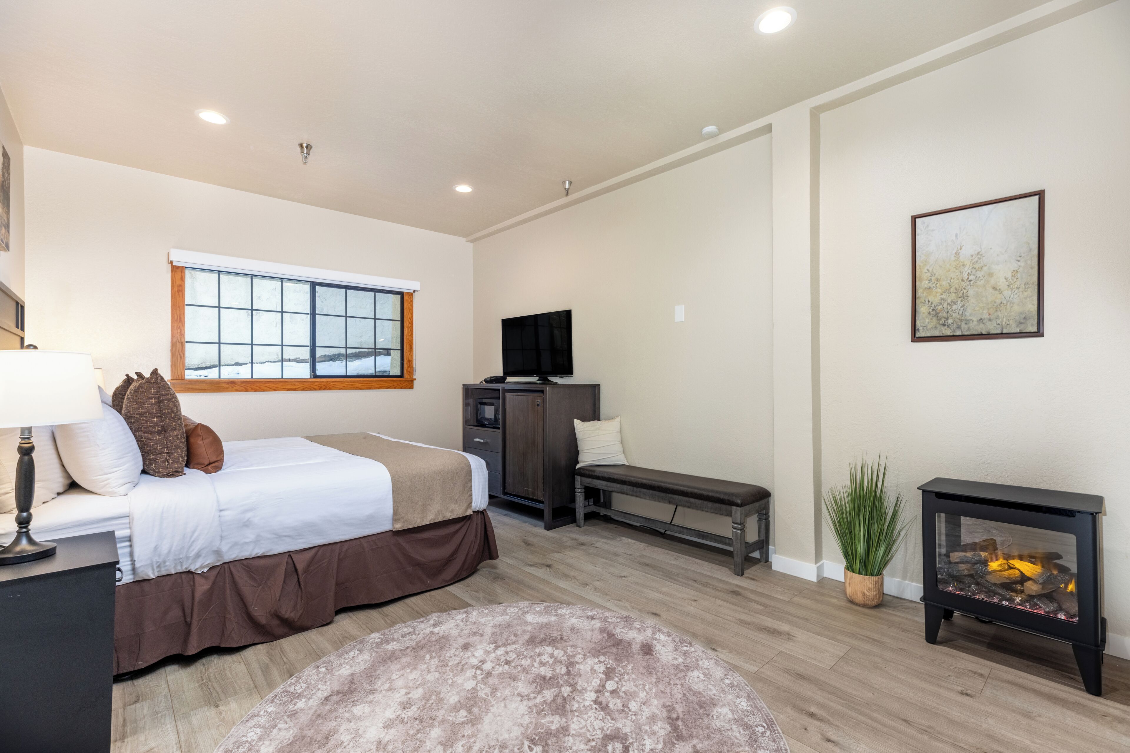 A bedroom with a bed, bedside table, lamp, TV, bench, and a fireplace.