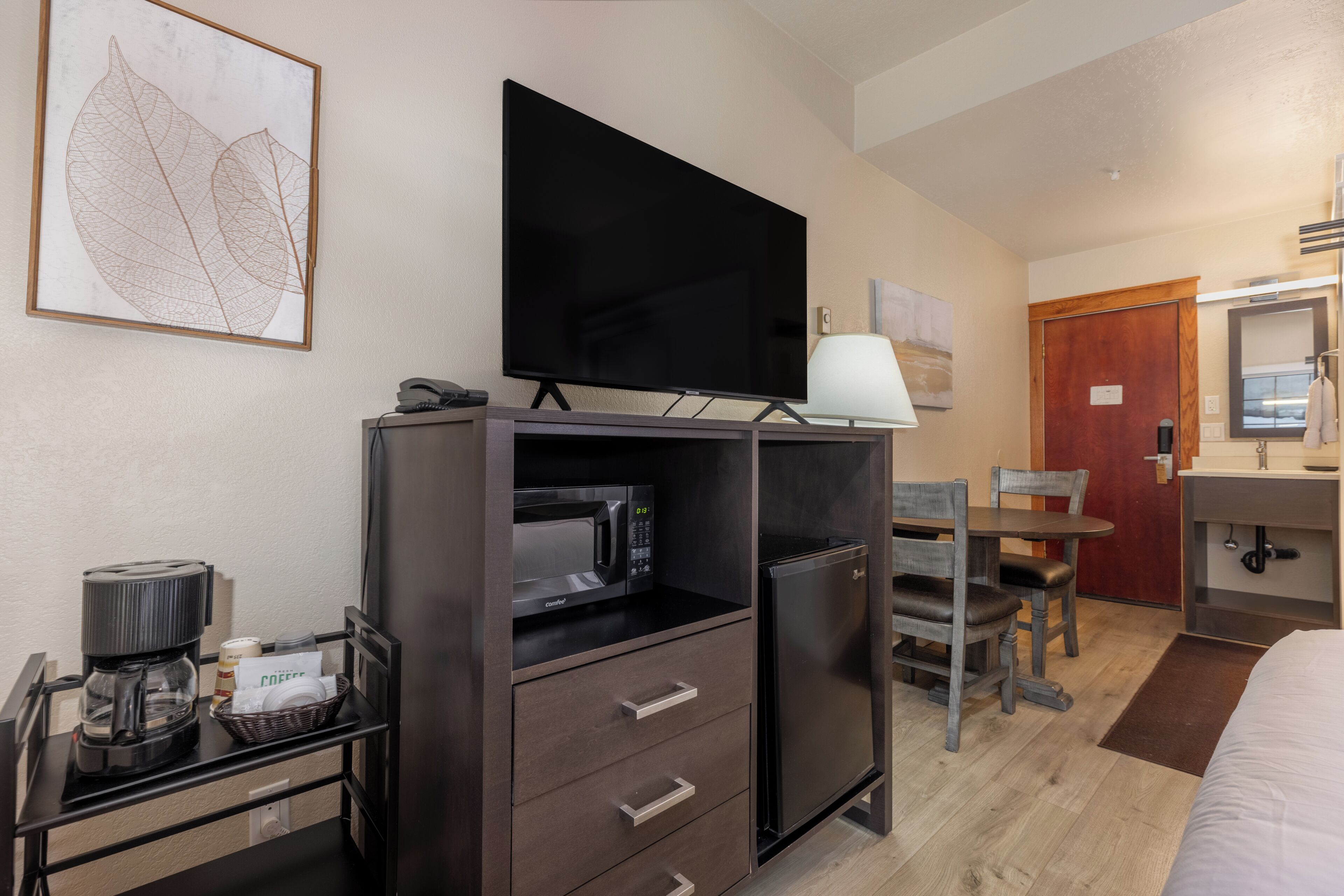 Quarto de hotel com televisão de tela plana, micro-ondas, geladeira, mesa de jantar com cadeiras, cama e banheiro com pia e espelho.