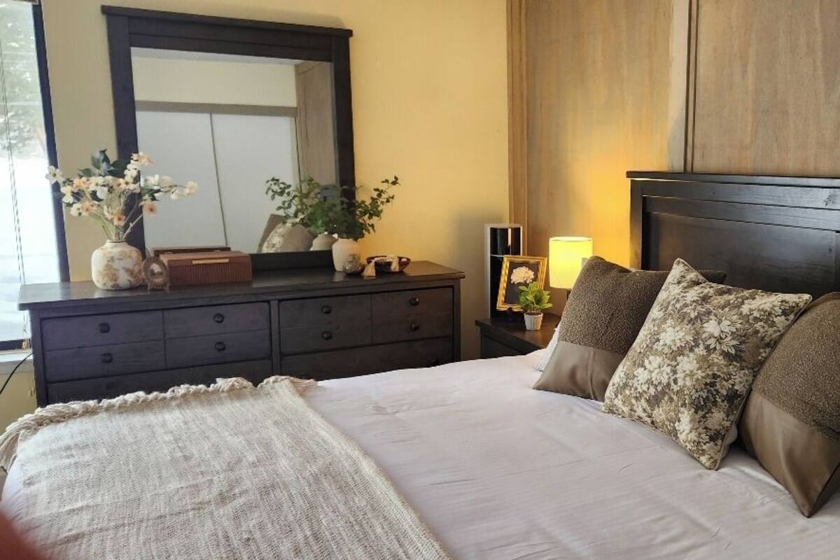 A bedroom with a large bed, dark wooden headboard, a dresser with drawers, a mirror, and a nightstand with a lamp.