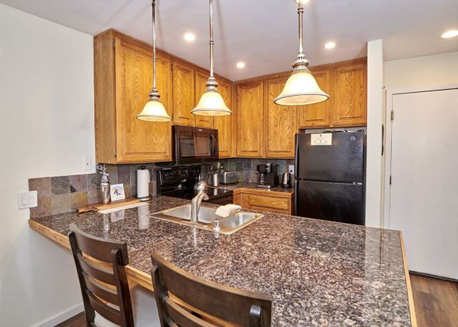 A kitchen with granite countertops, wooden cabinets, stainless steel appliances, and pendant lighting.