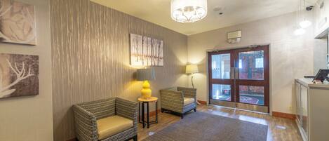 A hotel lobby with two wicker armchairs, a small table with a lamp, a patterned rug, a chandelier, and a door leading outside.