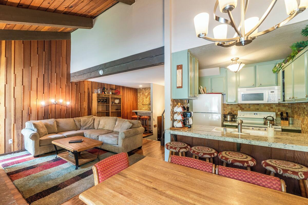 A modern living room with a sofa, coffee table, and ottomans, a kitchen with a microwave and stove, and a wooden ceiling.