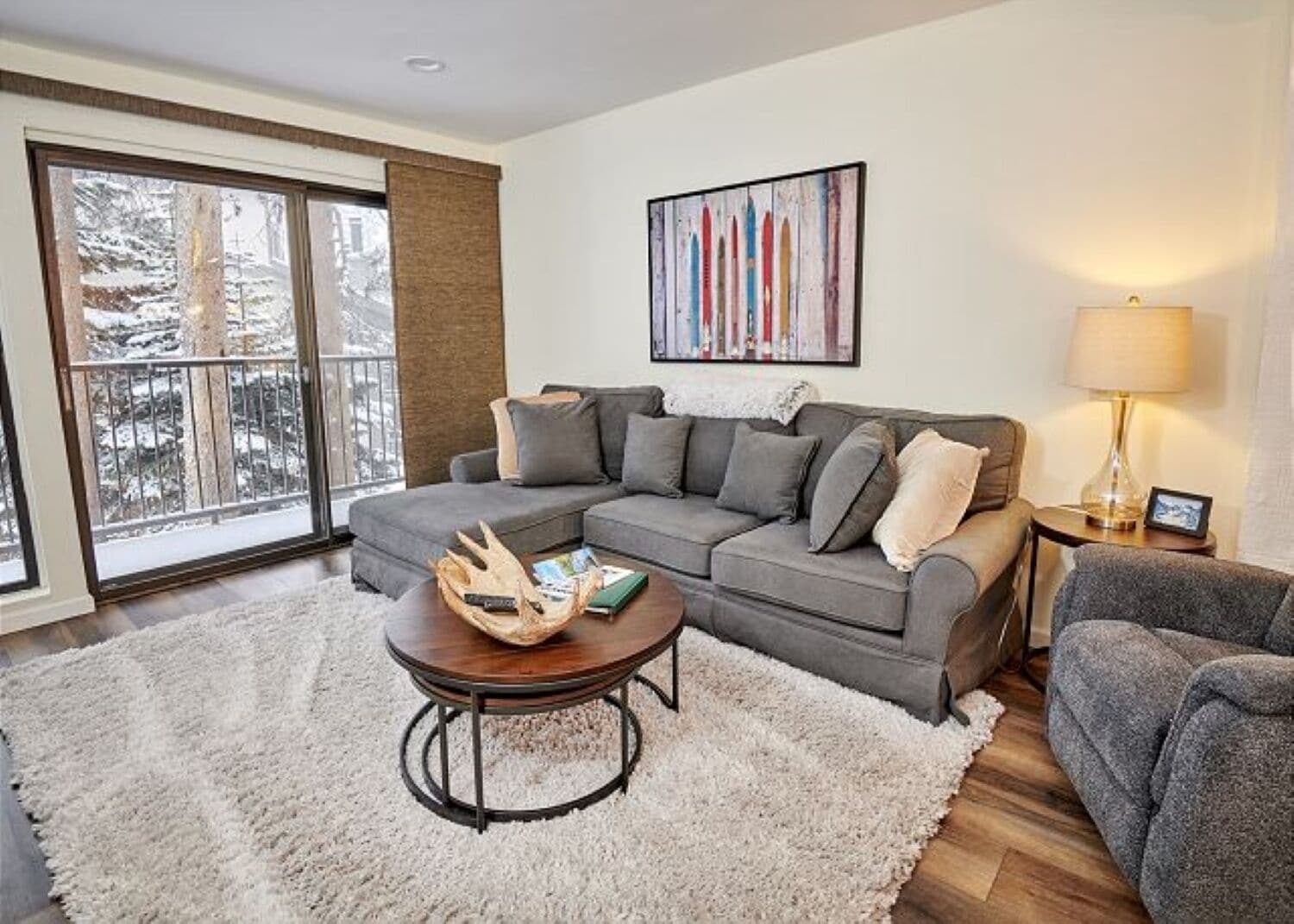A living room with a gray sectional sofa, a round coffee table, a white shag rug, a large window with a view of snow-covered trees, and a painting on the wall.