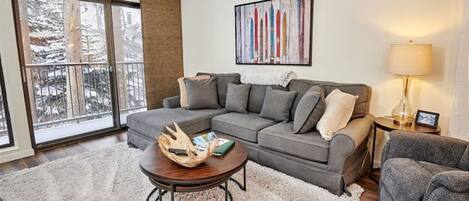 A living room with a gray sectional sofa, a round coffee table, a white shag rug, a large window with a view of snow-covered trees, and a painting on the wall.