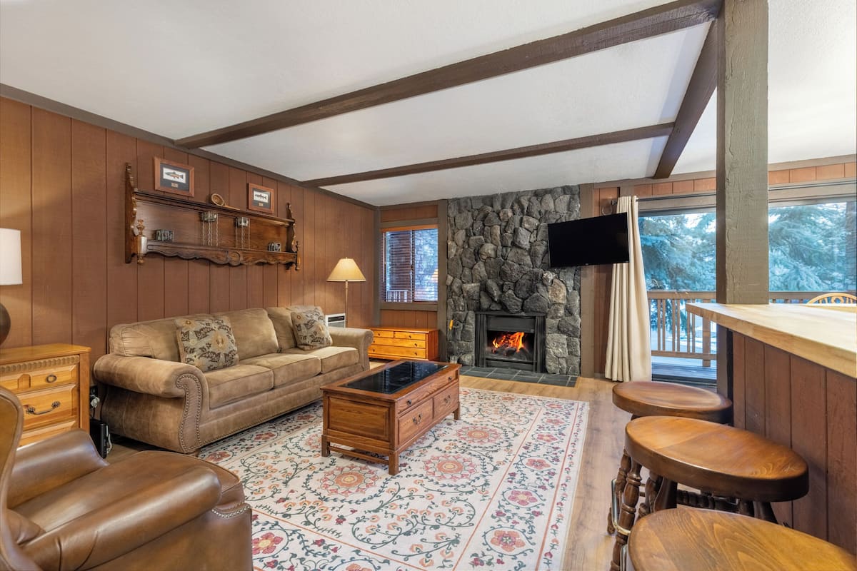 A cozy living room with a stone fireplace, wooden furniture, and a large window overlooking a snowy landscape.