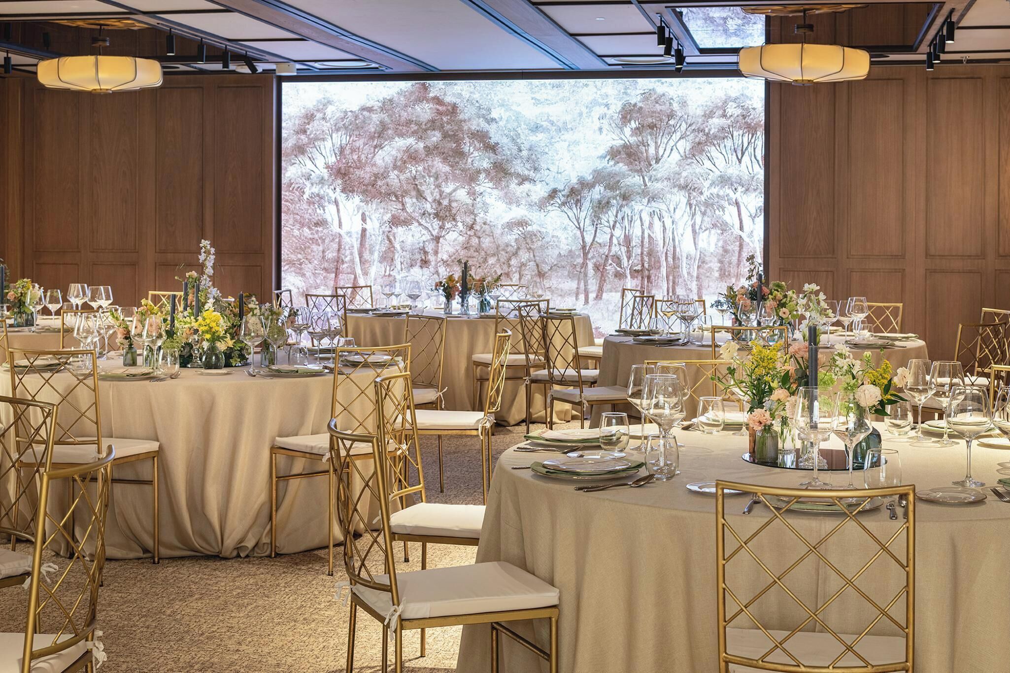 Un salón de banquetes con mesas redondas, sillas doradas y una gran pantalla de proyección que muestra una escena de bosque.