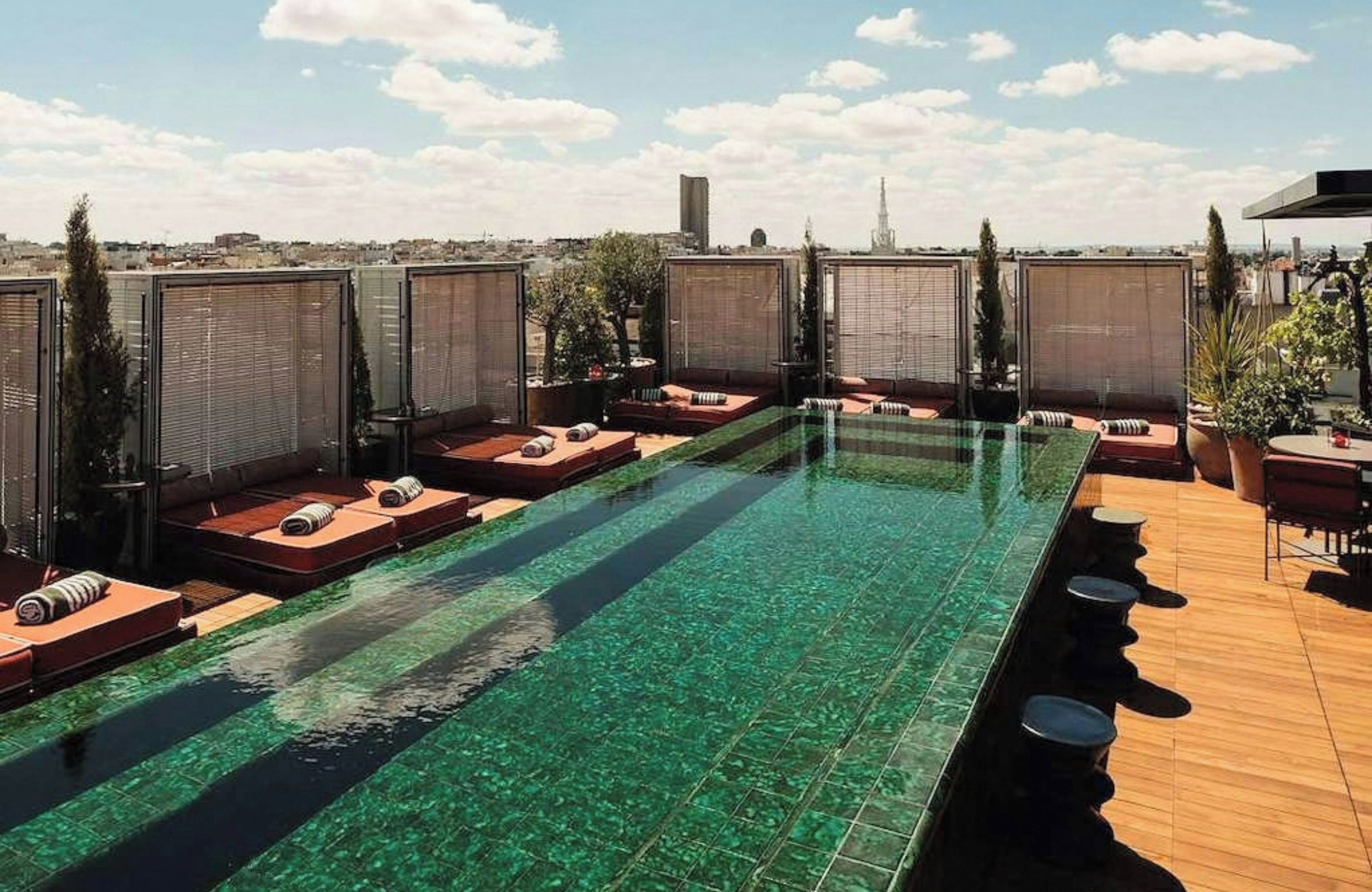 A rooftop pool with clear water, surrounded by lounge areas with sun loungers and potted plants.