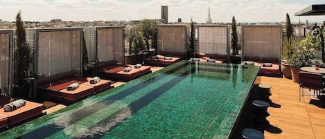A rooftop pool with clear water, surrounded by lounge areas with sun loungers and potted plants.