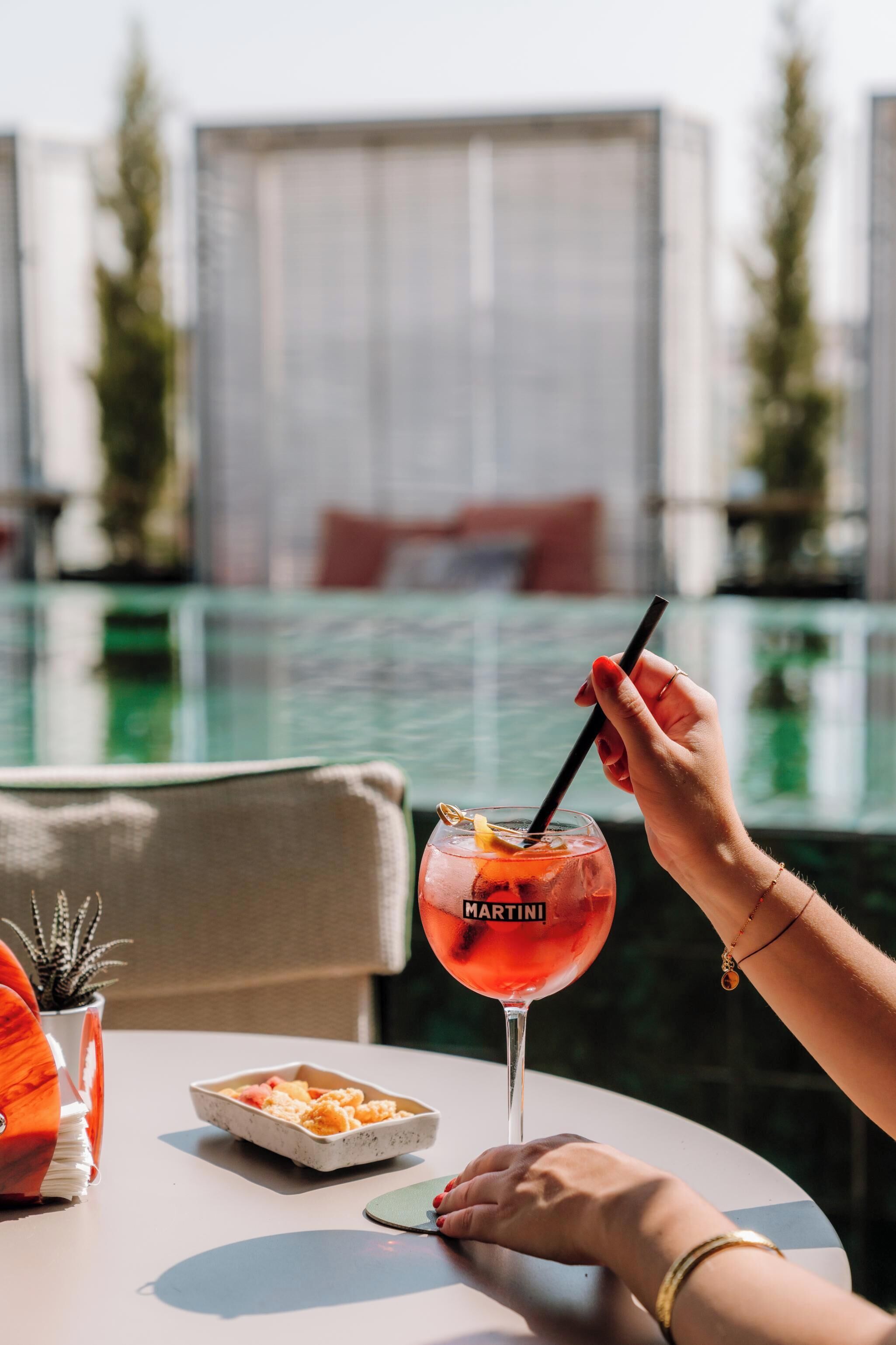 A person holding a Martini cocktail glass with a straw, a smartphone, and a small dish of food on a table.