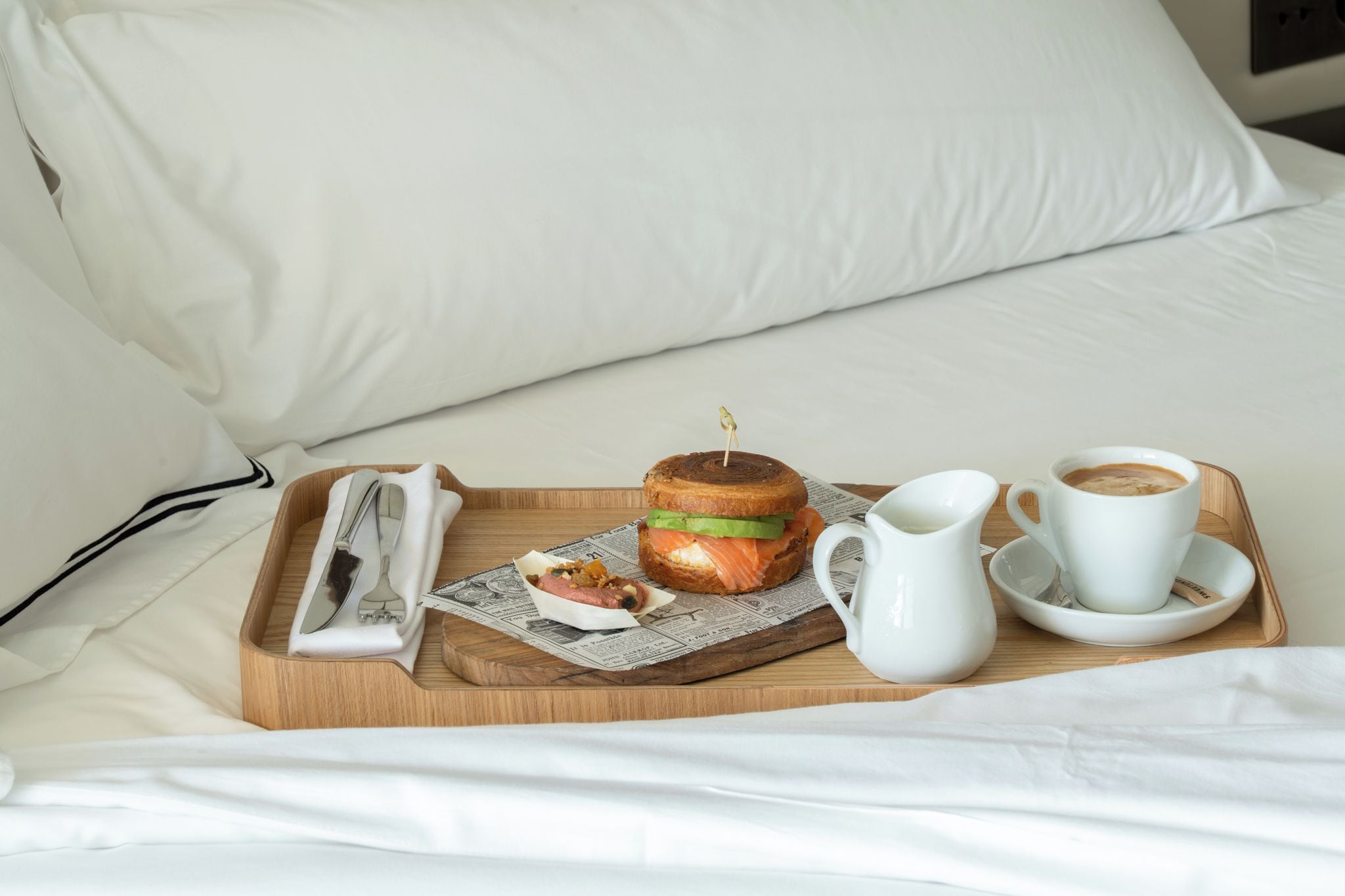 A wooden tray with a sandwich, a hot beverage, and a small plate with a fork and knife on a bed with white bedding.