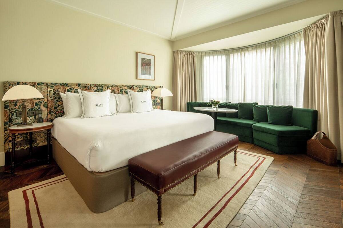 A hotel room with a large bed, a bench, a green sofa, a small table, and a window with curtains.