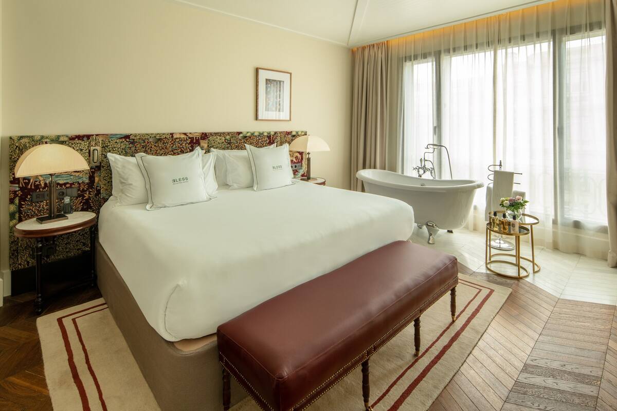 A hotel room with a large bed, a bench, a bathtub, and a small table with bottles.