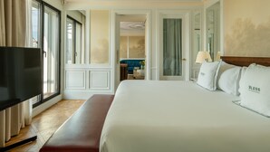 A hotel room with a large bed, a flat-screen TV, a bench, and a view of the city through large windows.