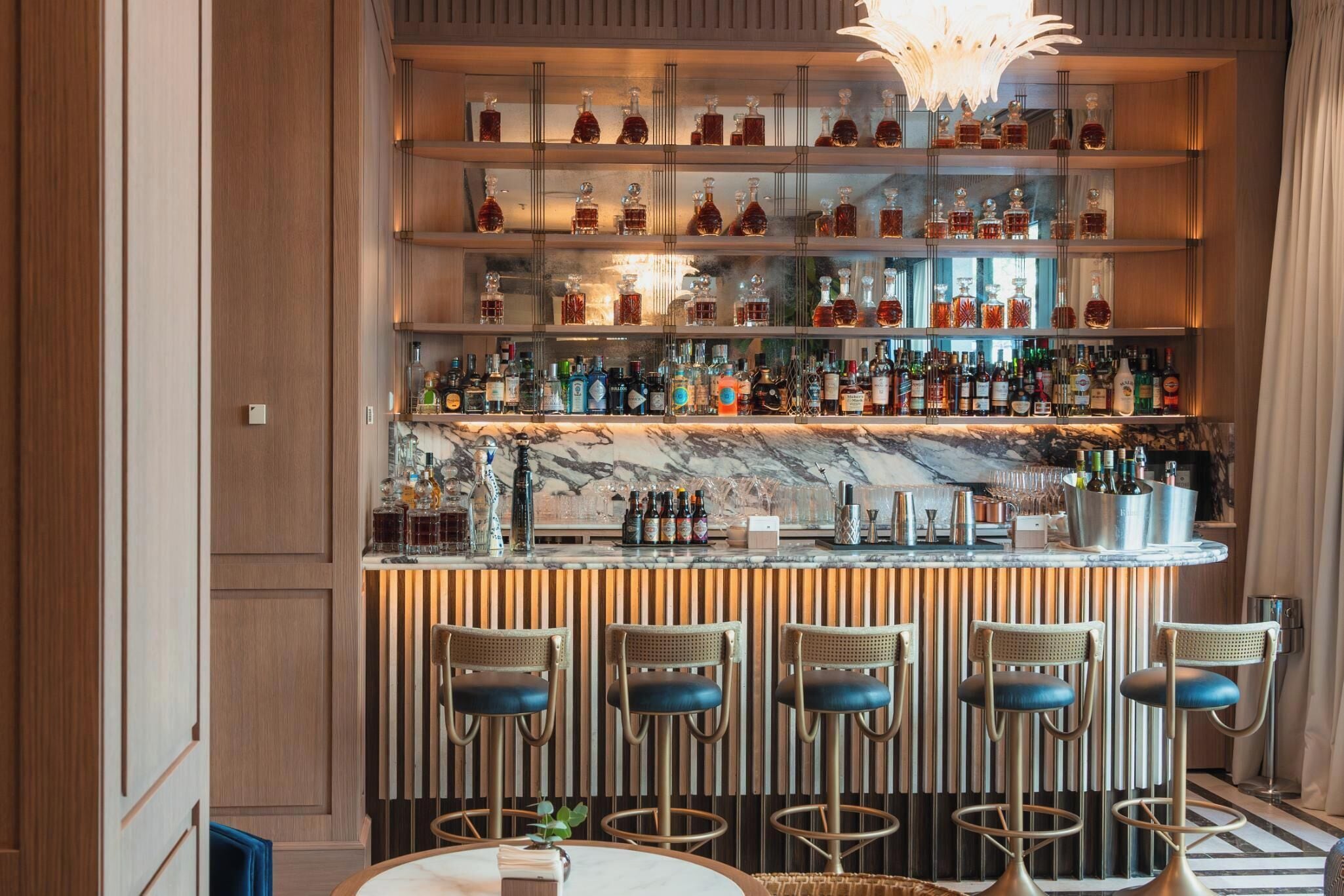 Zona de bar con varias estanterías llenas de botellas de licor, una encimera de mármol y taburetes.