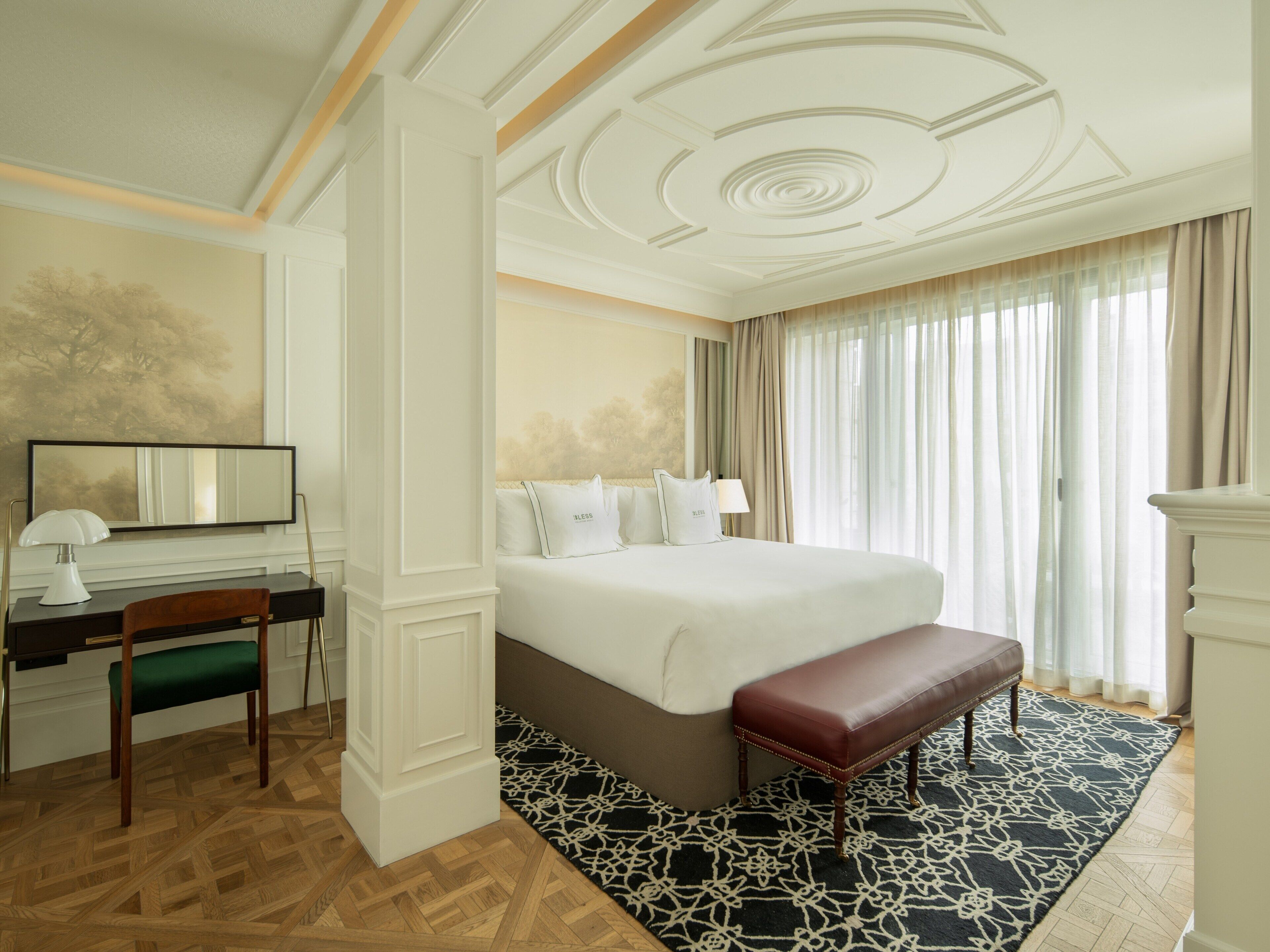 A hotel room with a large bed, a desk with a chair, a bench, a large mirror, and a patterned rug.