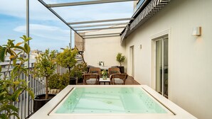 A rooftop terrace with a glass-enclosed hot tub, wicker furniture, potted plants, and a view of the cityscape.