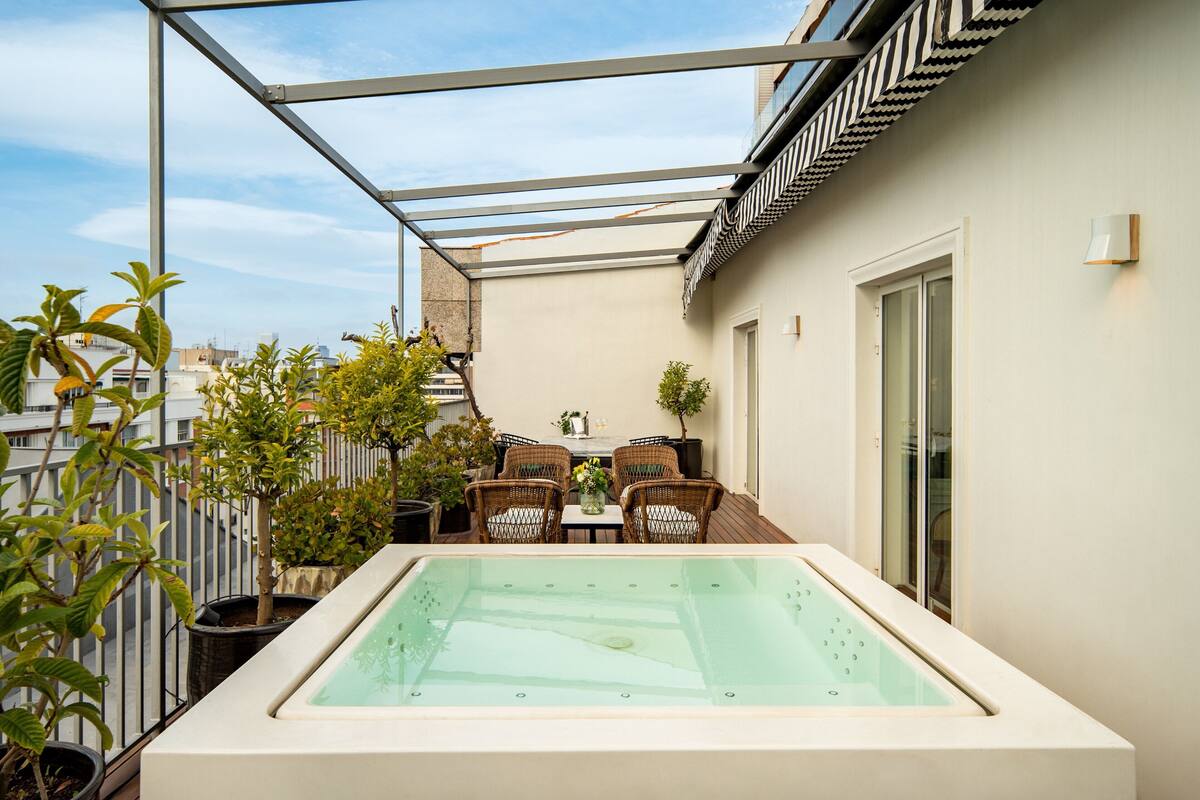 A rooftop terrace with a glass-enclosed hot tub, wicker furniture, potted plants, and a view of the cityscape.