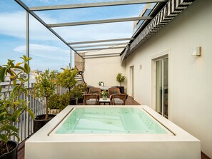 Een terras op het dak met een glazen hottub, rieten meubels, potplanten en uitzicht op de skyline.