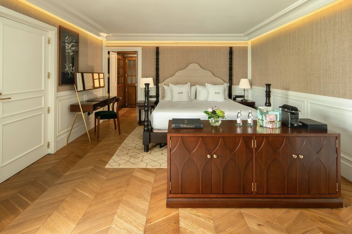 A hotel room with a large bed, a wooden dresser, a desk with a chair, and a small table with a vase of flowers.