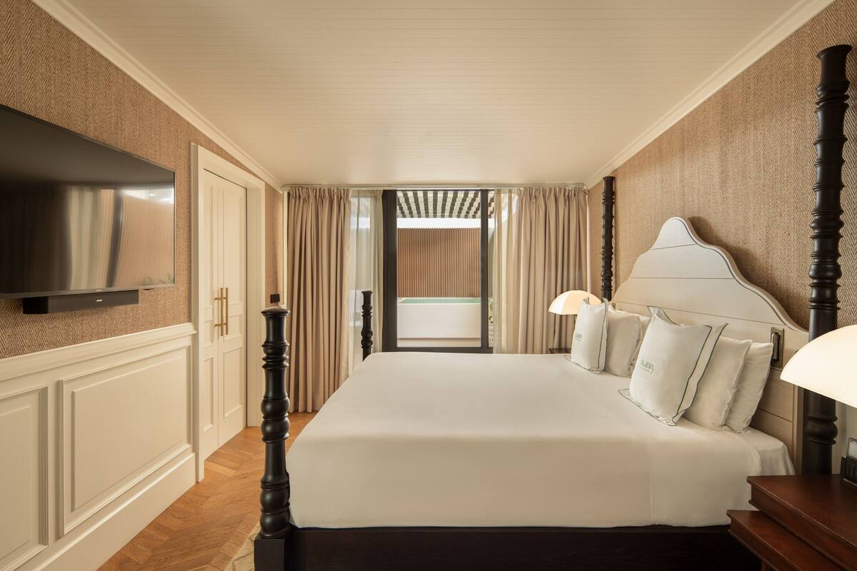 A hotel room with a large bed, a flat-screen TV mounted on the wall, a wooden nightstand, and a window with curtains.