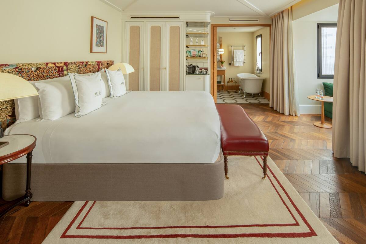 A hotel room with a large bed, a bench, a desk, a chair, a bathroom, and a view of the city.