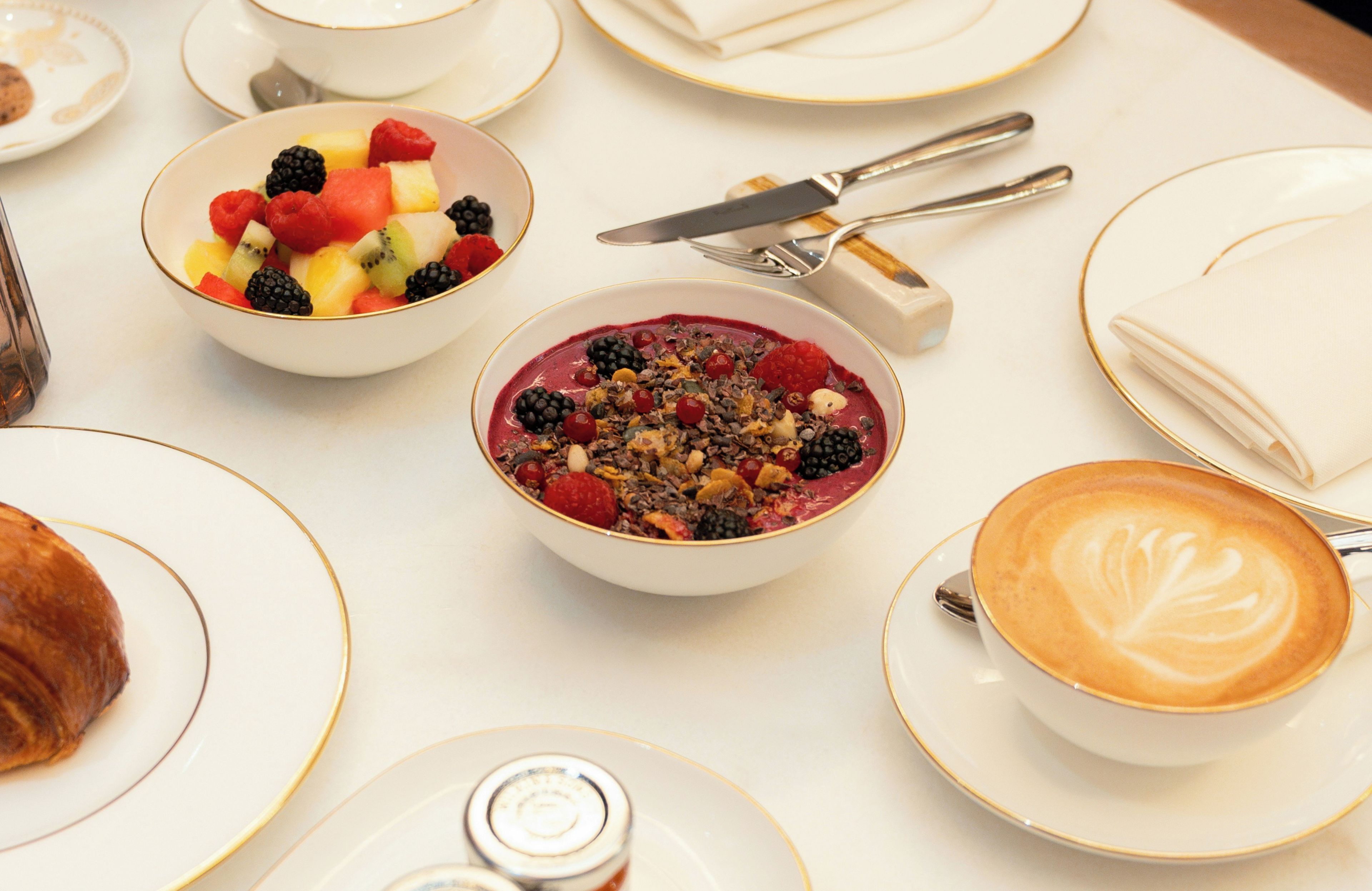 Una mesa con distintos productos de desayuno, incluyendo cuencos de fruta, una taza de café con arte de latte, y un juego de cuchillo y tenedor.