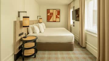 A hotel room with a large bed, a small round table with chairs, a wall-mounted TV, and a large window with curtains.