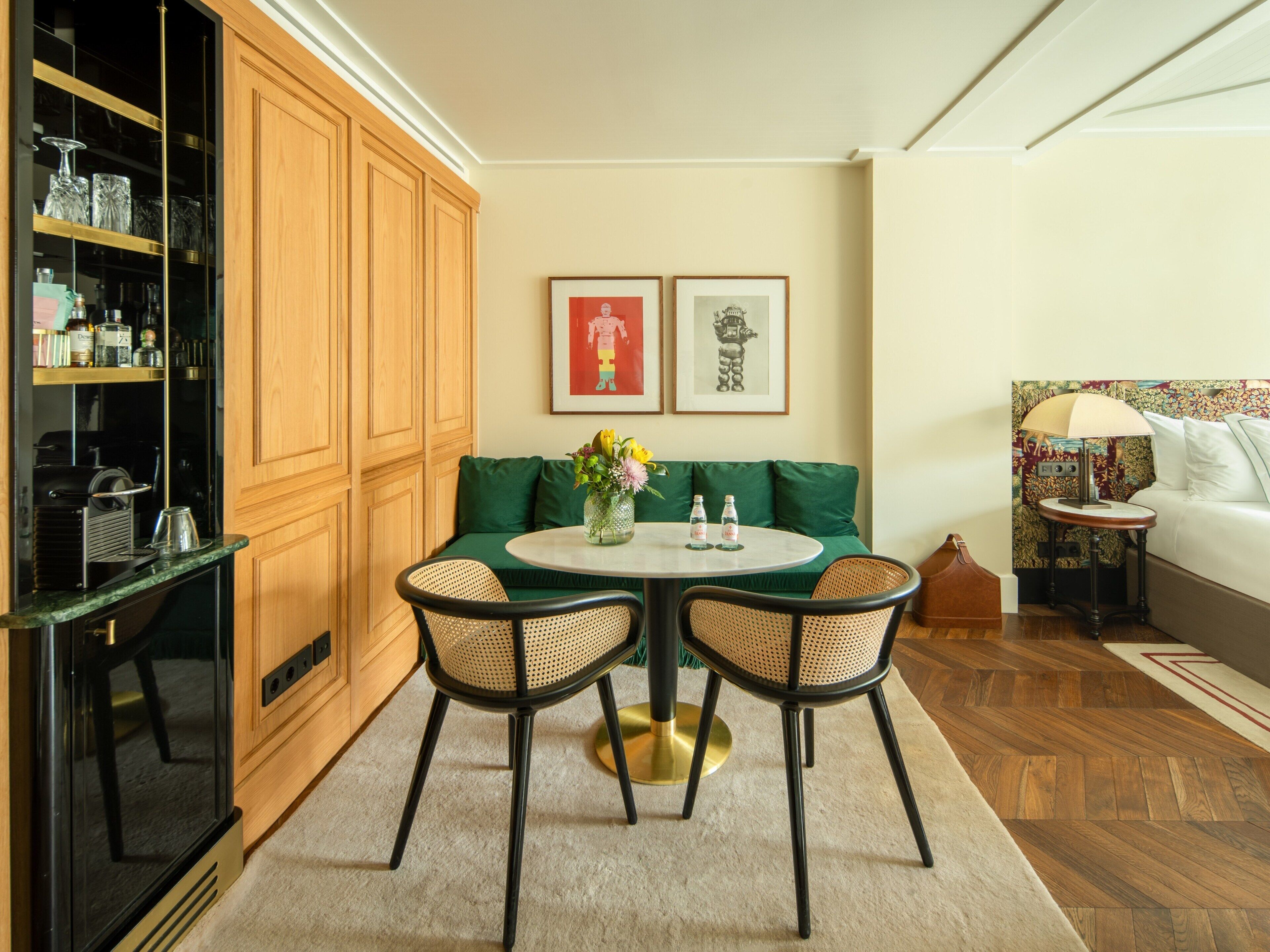 A cozy living room with a green sofa, two wicker chairs, a round table with a vase and bottles, and a cabinet with glassware.