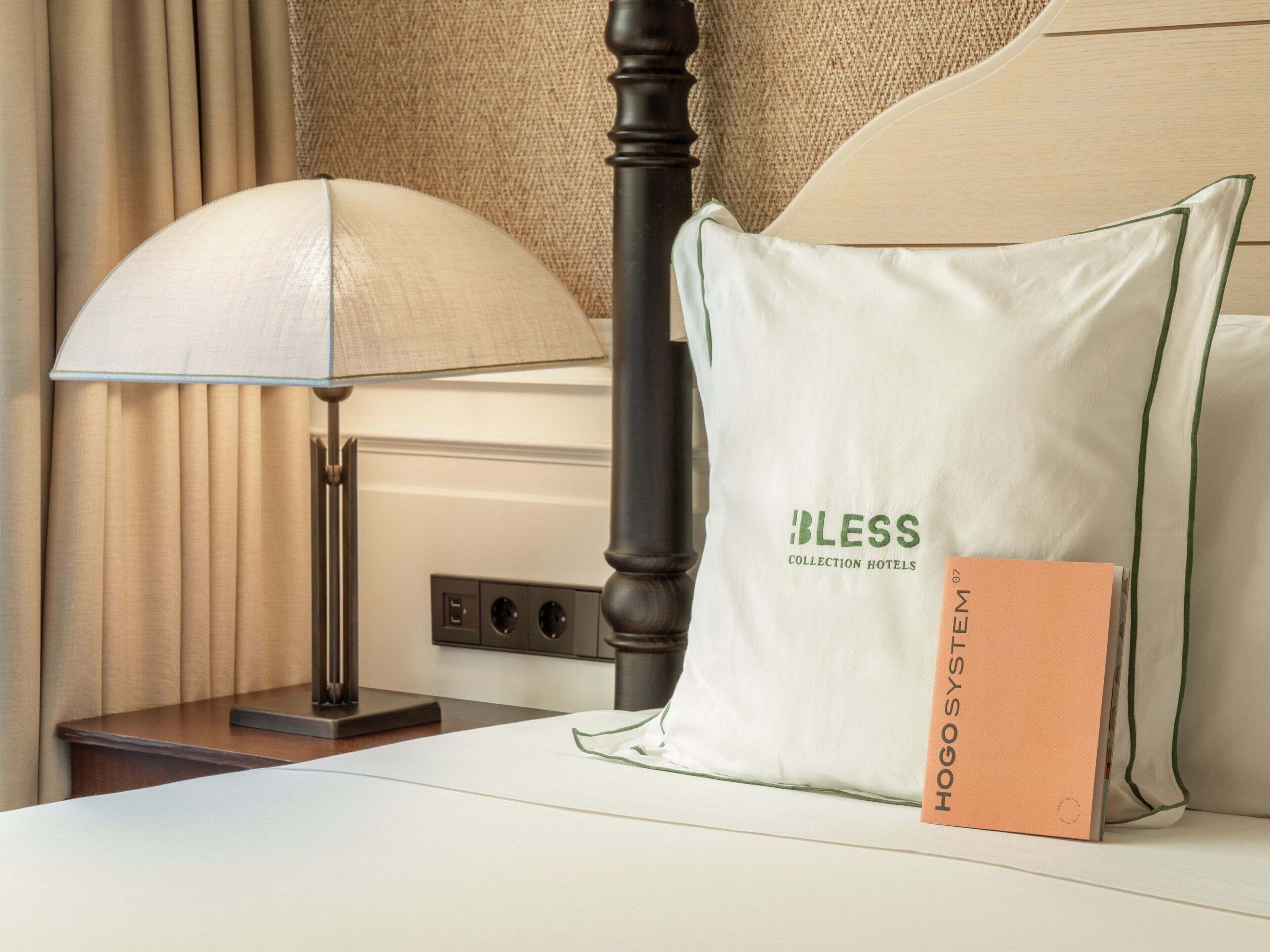 Una cama de hotel impecable con un cojín que dice 'Bless Collection Hotels' y un folleto titulado 'Hogsty System'.