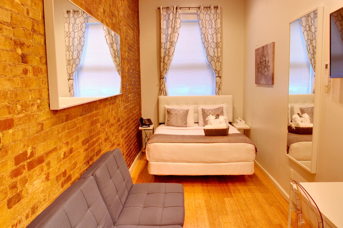 A hotel room with a brick wall, a large bed with white bedding, a grey sofa, and a small bedside table.