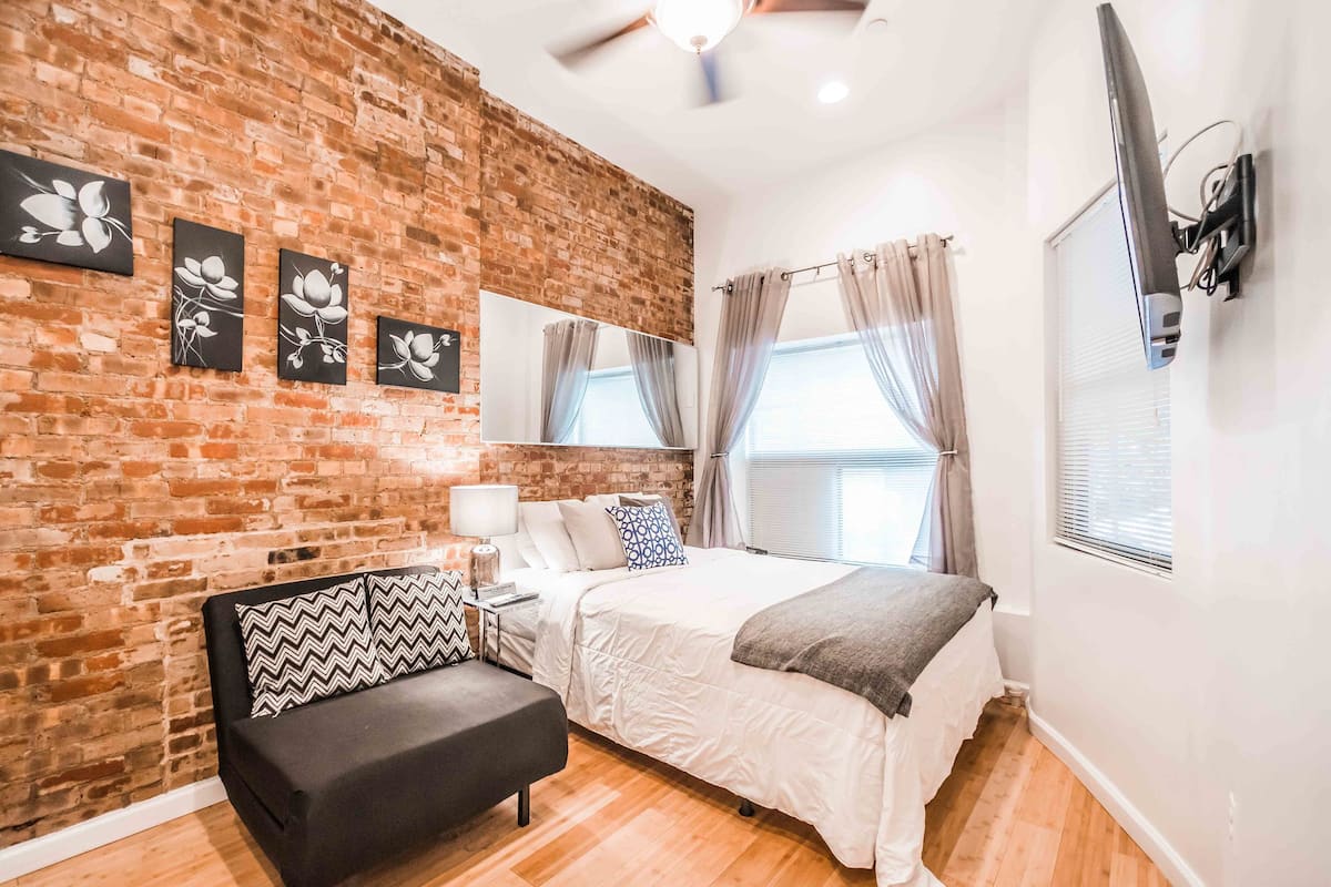 A bedroom with a brick wall, a bed with white bedding, a black sofa with patterned cushions, a bedside table, a mirror, and a ceiling fan.