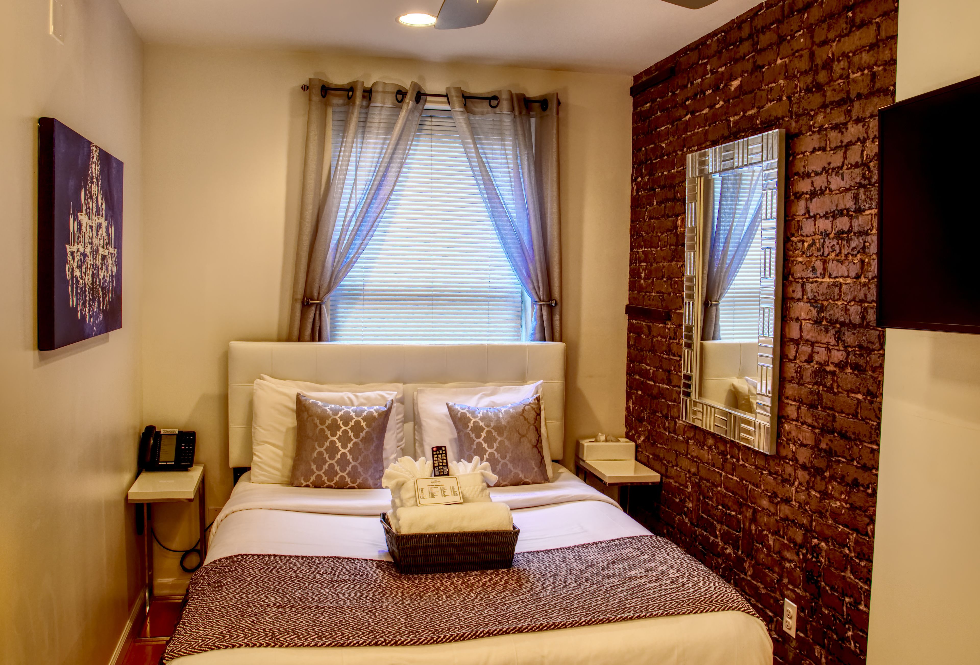 A hotel room with a brick wall, a large bed with white bedding, a bedside table with a lamp, and a window with curtains.