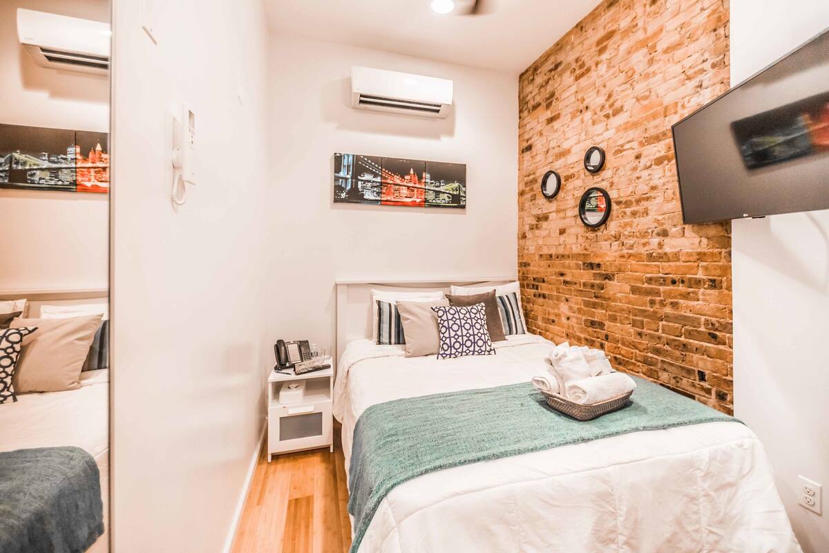A bedroom with a brick accent wall, a bed with a green throw, a bedside table with a phone and a lamp, and a TV mounted on the wall.