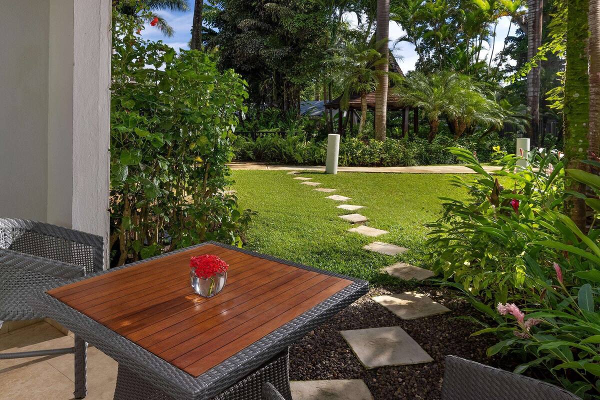 Un patio con una mesa de madera y sillas de mimbre, rodeado de abundante vegetación y un sendero de piedra.