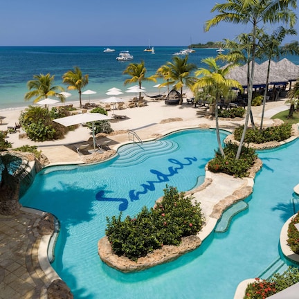 A resort pool with 'Sandals' written in the water, surrounded by palm trees, beach chairs, and umbrellas.
