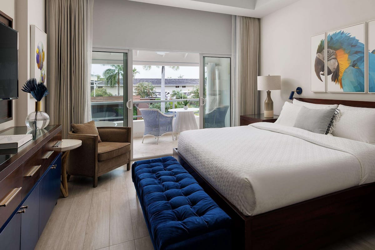 Une chambre d’hôtel dotée d’un grand lit, d’un coin salon avec un canapé et des fauteuils, d’un balcon offrant une vue sur les palmiers et d’œuvres d’art accrochées aux murs.