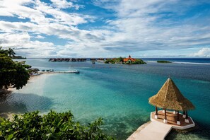 Une plage tropicale aux eaux turquoise et limpides, un gazebo à toit de chaume et des bungalows sur pilotis.