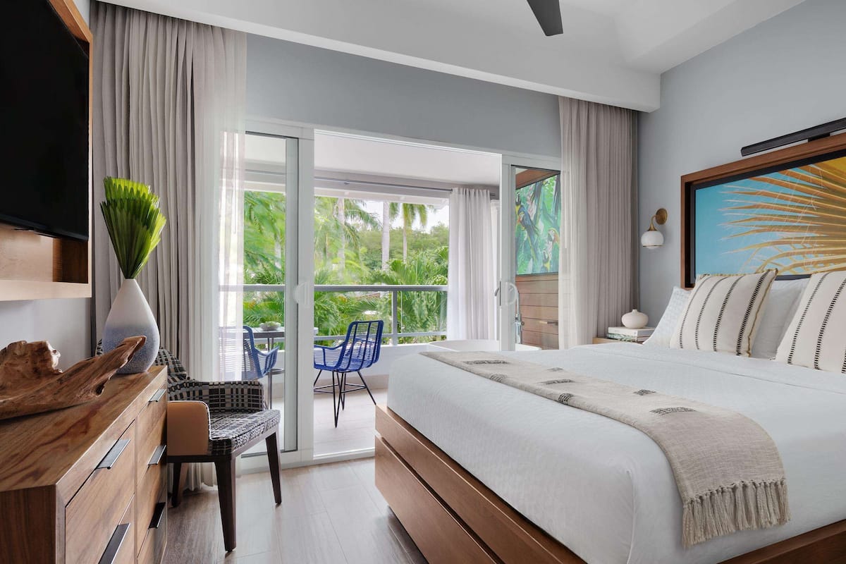 Une chambre moderne avec un grand lit, un bureau et une chaise. Elle dispose d’un balcon avec vue sur la verdure.