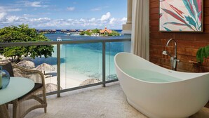 Un baño moderno con una bañera independiente, un balcón acristalado con vistas a una playa tropical y bungalows sobre el agua.